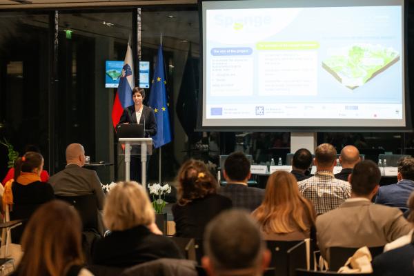 A speaker presents a project on a screen during an event, with participants seated in front of her. Flags of Slovenia and the European Union are visible in the background.